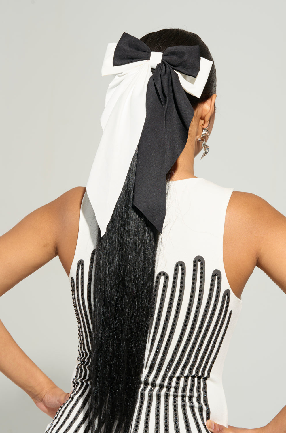 A woman facing away from the camera wears a sleeveless dress with a colorblock design accented by black and white detailing. Her long black hair is elegantly tied back using the SPLIT DECISION HAIR BOW. She stands with her hands on her hips, wearing star-shaped earrings that add a whimsical touch to her outfit. 1