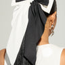 A person with long black hair adorned by the SPLIT DECISION HAIR BOW, a large accessory in black and white. They sport silver dangling earrings and a white top featuring an oversized silhouette enhanced with black beads. The background is plain and light-colored.