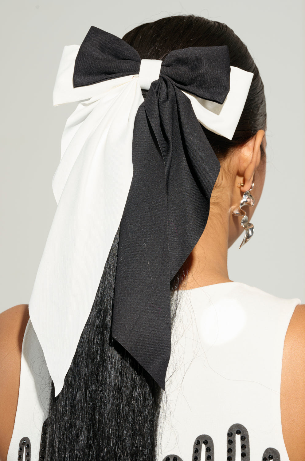 A person with long black hair adorned by the SPLIT DECISION HAIR BOW, a large accessory in black and white. They sport silver dangling earrings and a white top featuring an oversized silhouette enhanced with black beads. The background is plain and light-colored. 0