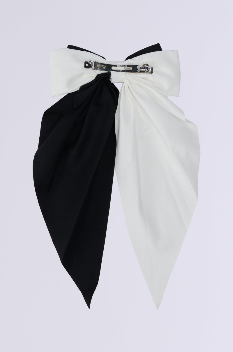 The SPLIT DECISION HAIR BOW showcases a colorblock design with its oversized silhouette, featuring one half in black and the other in white, joined by a central silver clasp. Its long, cascading tails stand out against the plain gray background. 3