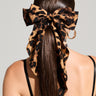 A woman with long, straight brown hair showcases a MILANO CHEETAH HAIR BOW tied at the back of her head. She is wearing a black spaghetti-strap top and hoop earrings are noticeable. The background remains simple and unobtrusive.