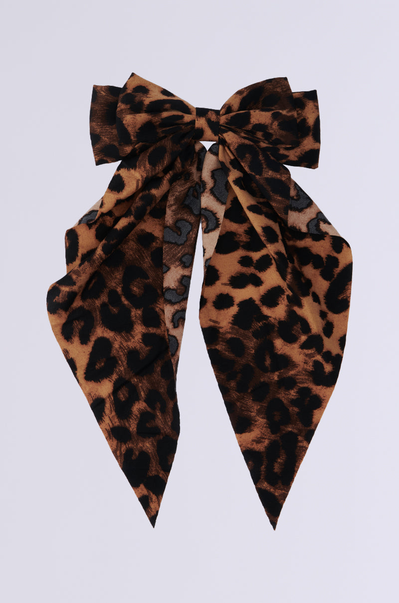 The MILANO CHEETAH HAIR BOW is an oversized accessory showcasing a striking leopard print with brown and black spots set on a light gray background. Its long, pointed tails drape elegantly across a plain white surface. 1