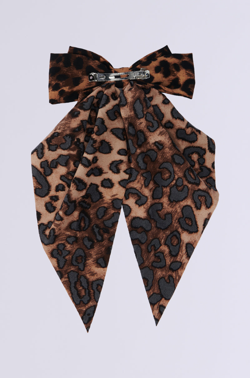 The MILANO CHEETAH HAIR BOW is an oversized accessory featuring a large bow on top with two long, pointed tails. It showcases a stylish blend of brown and black spots and includes a metal clasp for easy attachment. 2