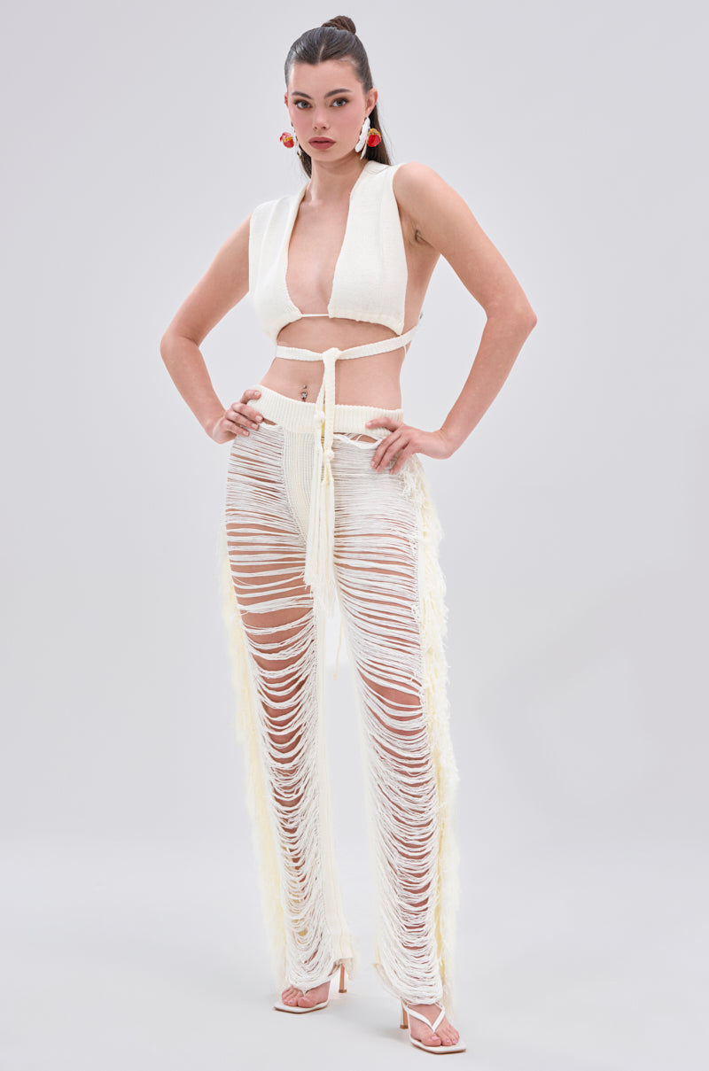 A woman poses confidently in the BEST DRESSED FRINGE BRALETTE AND LEGGING, featuring a white cropped top with a tie detail and sheer, shredded leggings. She completes the look with high heels, red earrings, and her hair tied back.