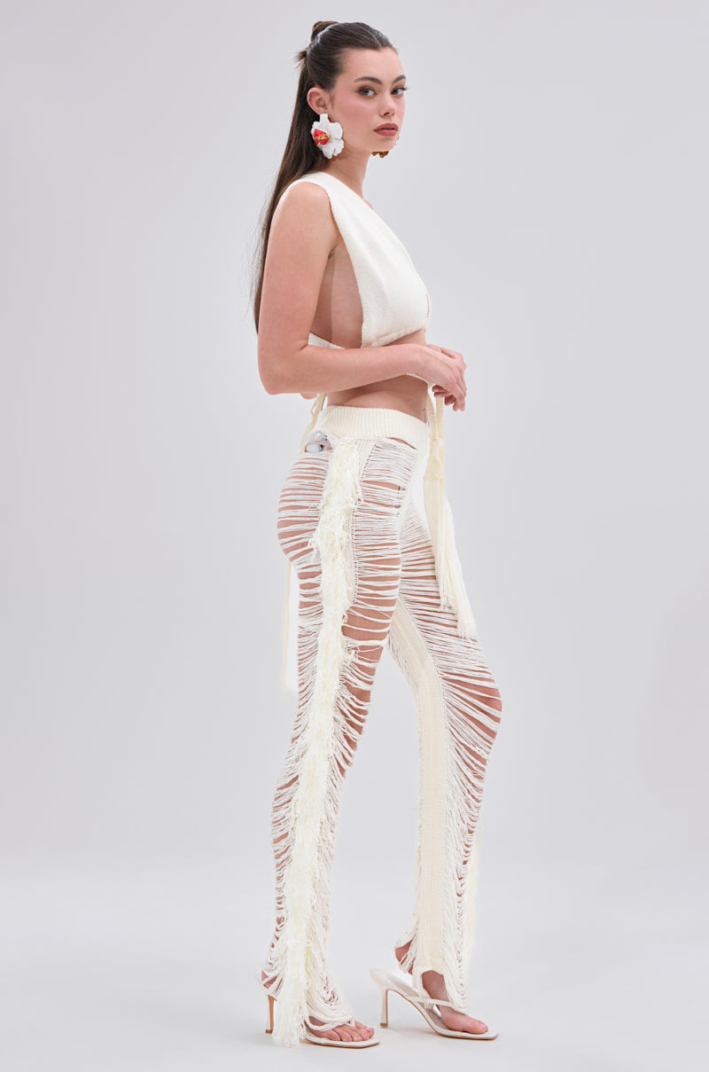 A woman stands sideways in the BEST DRESSED FRINGE BRALETTE AND LEGGING set— a sleeveless white top and fringed, cut-out pants with an elastic waistband. She wears high heels, has her long hair tied back, and accessorizes with a white flower earring. Light gray background.