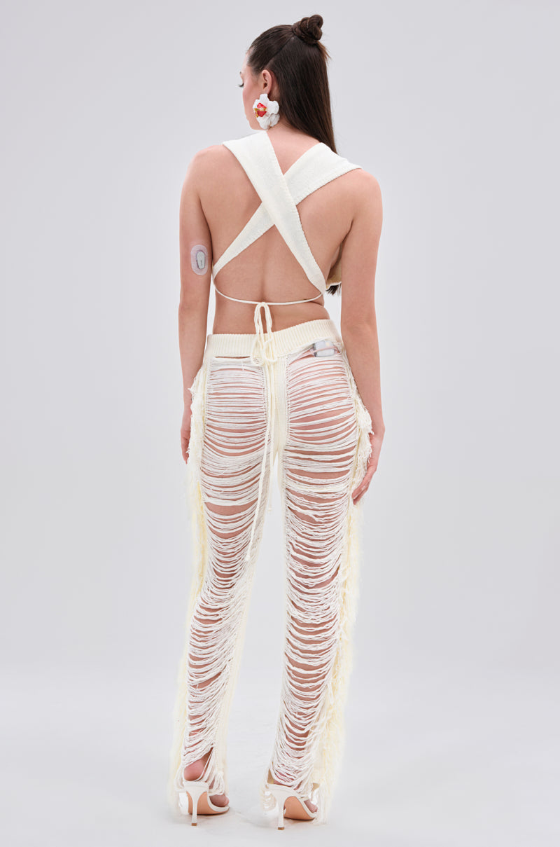 A person stands with their back to the camera, wearing the BEST DRESSED FRINGE BRALETTE AND LEGGING set, white heeled sandals, large earrings, and an adhesive medical patch on their upper arm against a plain light gray background.