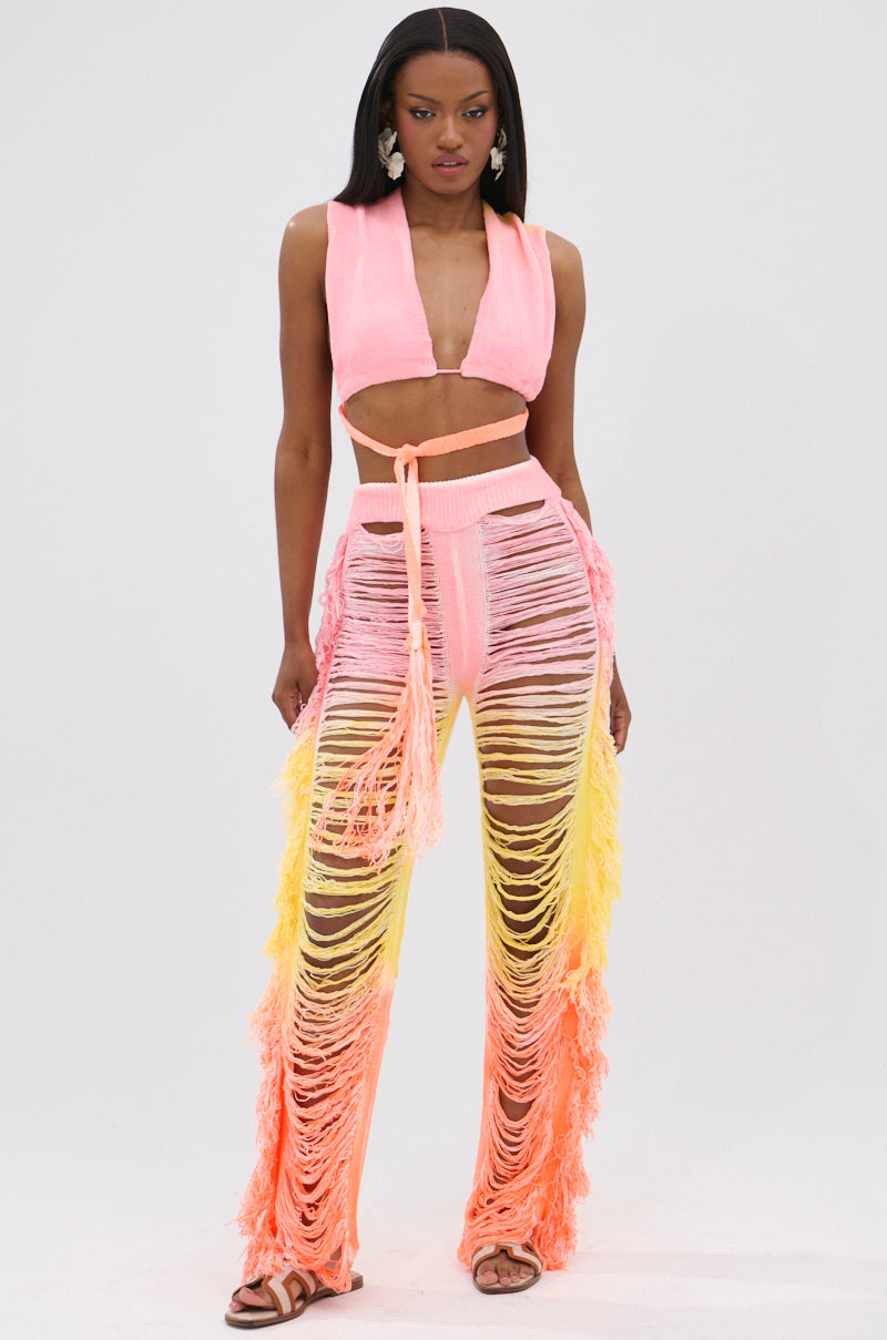 A woman faces forward in the TIME TO DANCE CUTOUT FRINGE PANT AND TOP SET, featuring a pink convertible top and ombré high-waisted cutout fringe pants in pink, yellow, and orange. She has straight hair, sandals, and stands against a plain background.