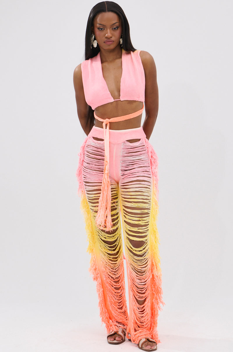 A woman with long, straight hair and a serious expression models the TIME TO DANCE CUTOUT FRINGE PANT AND TOP SET—pink convertible crop top and high-waisted pants with yellow and pink fringed cutouts—styled with sandals and statement earrings.