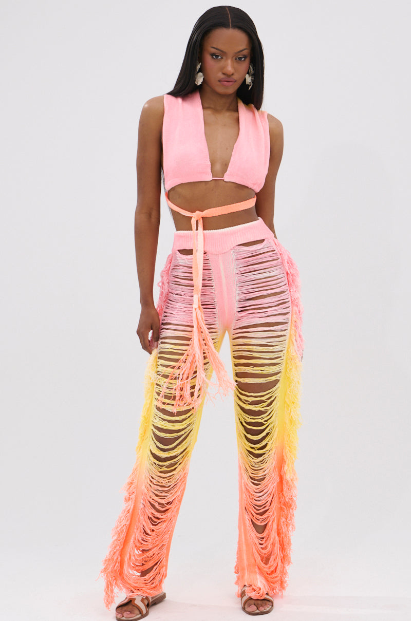 A woman poses in the TIME TO DANCE CUTOUT FRINGE PANT AND TOP SET, featuring a sleeveless convertible top and high-waisted pants with pink, yellow, and orange gradient fringe. She completes the vibrant look with sandals and statement earrings.