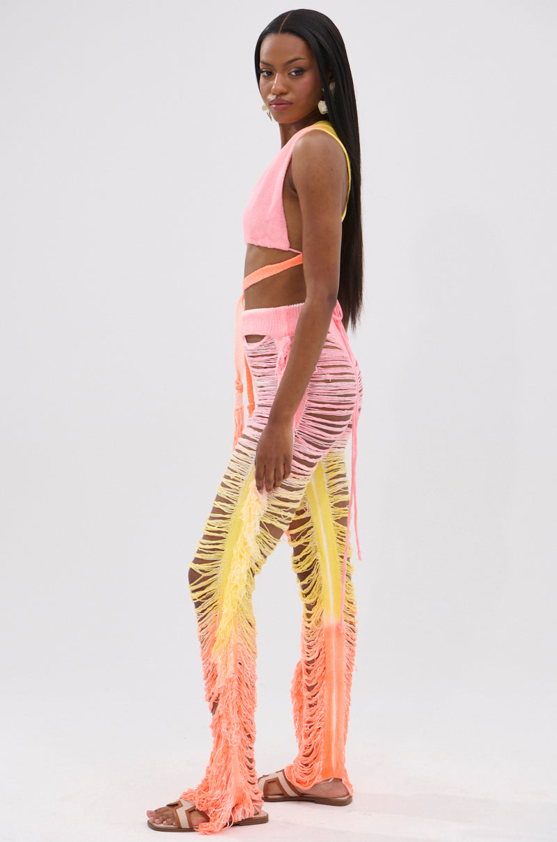 A woman stands sideways in the TIME TO DANCE CUTOUT FRINGE PANT AND TOP SET, featuring a sleeveless convertible top and matching high-waisted cutout fringe pants with bold pink and yellow shredding. She wears flat sandals and has long straight hair.