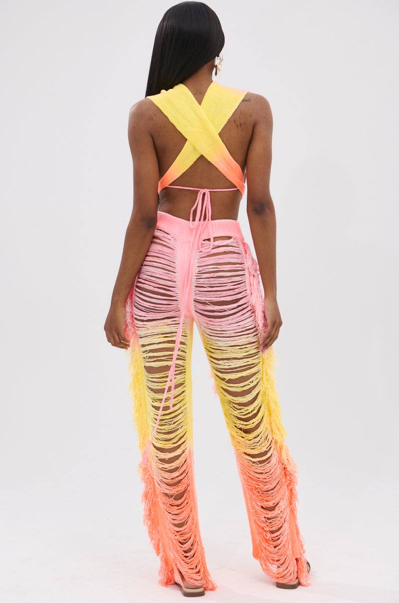 A person faces away, wearing the TIME TO DANCE CUTOUT FRINGE PANT AND TOP SET in yellow, pink, and orange. The vibrant high-waisted pants with shredded details make this look perfect for a lively summer soirée.
