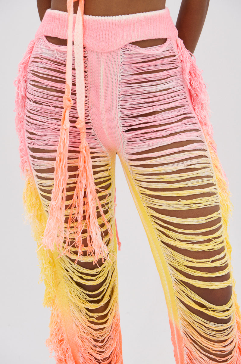 A close-up of a person wearing the TIME TO DANCE CUTOUT FRINGE PANT AND TOP SET, featuring high-waisted pink, yellow, and orange gradient pants with long tassels, cutouts, and horizontal slits revealing skin.