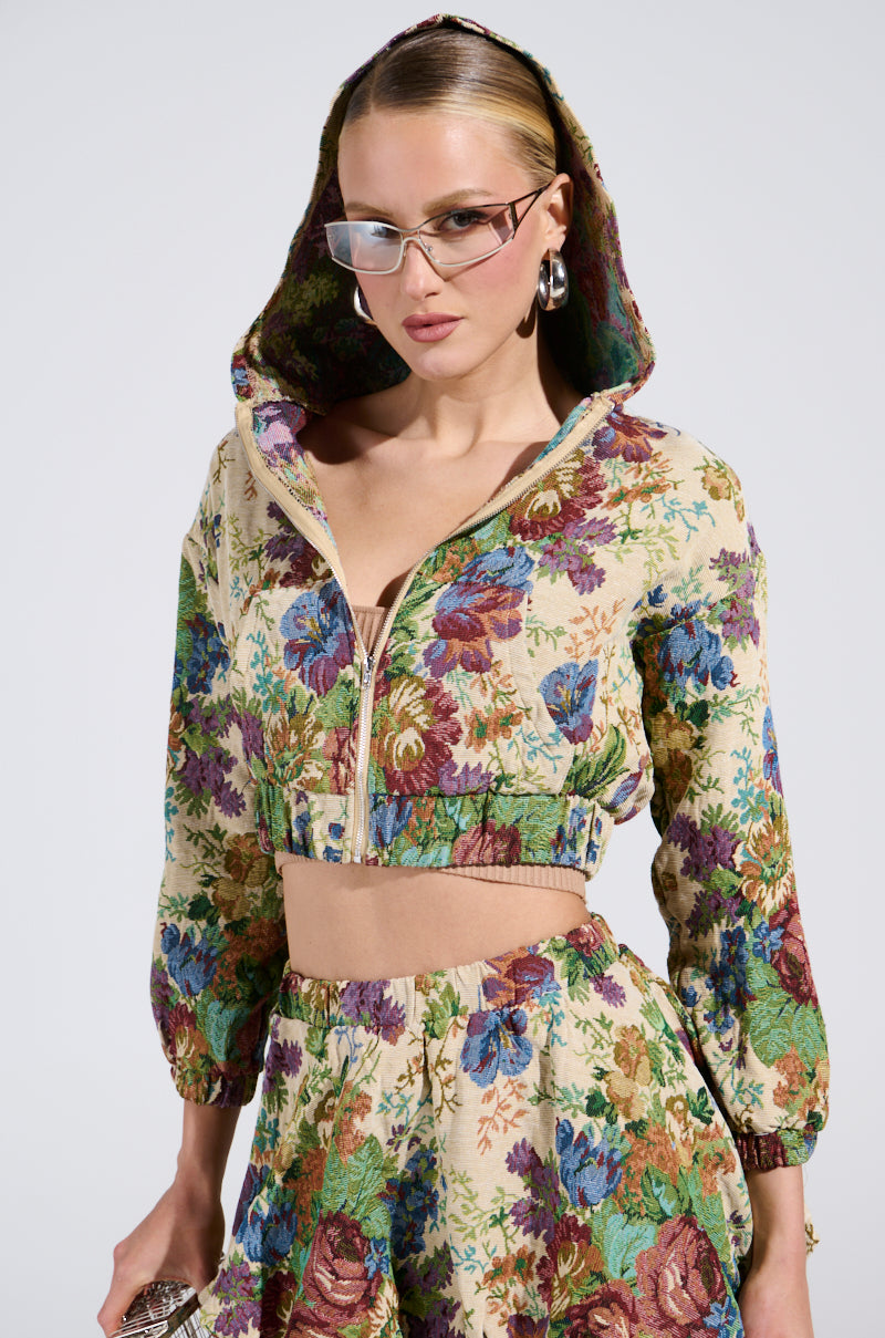 A person confidently wears a vibrant ALL ABOUT ME FLORAL BOMBER JACKET and matching pants, accessorized with stylish glasses, large hoop earrings, and a small purse. The ensemble exudes vintage charm with colorful flowers on a light background.