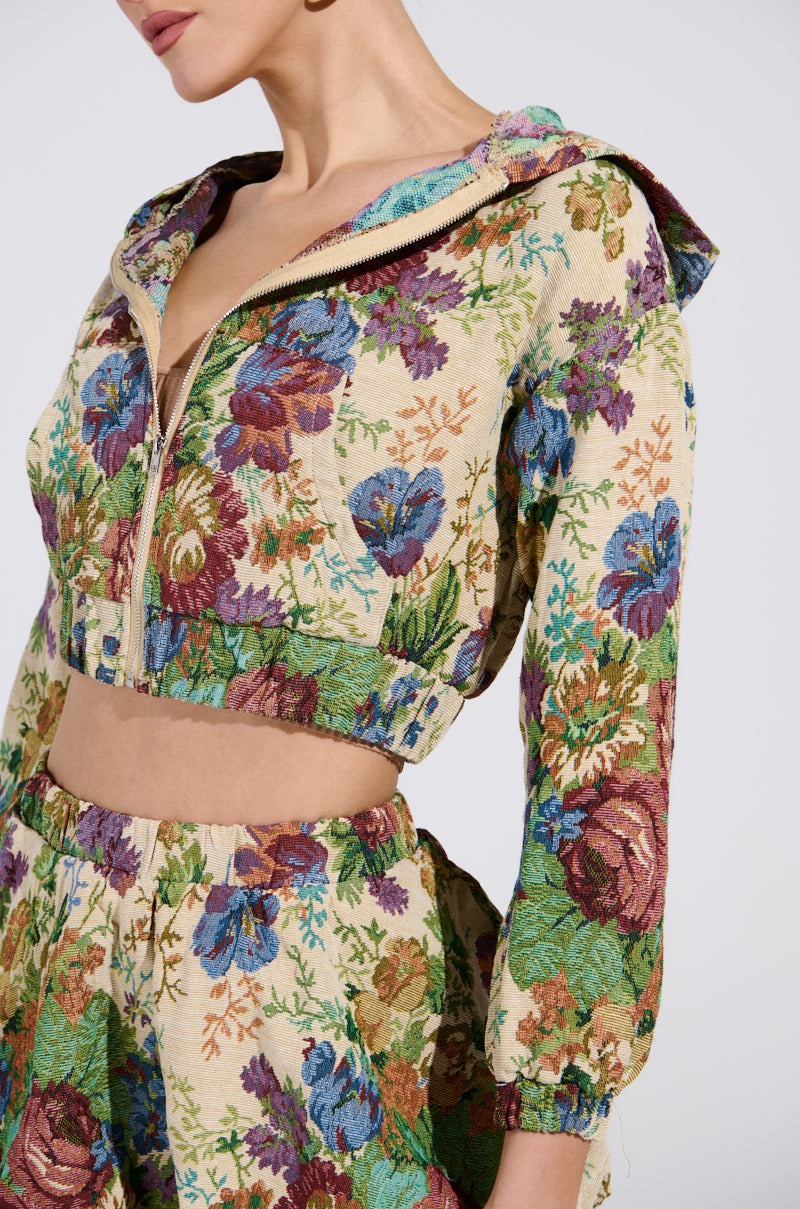 Dressed in the ALL ABOUT ME FLORAL BOMBER JACKET and matching high-waisted shorts, a person exudes vintage charm with vibrant blue, purple, and pink flowers on beige. They pose against a light gray background, capturing the AKIRA Label's signature style.