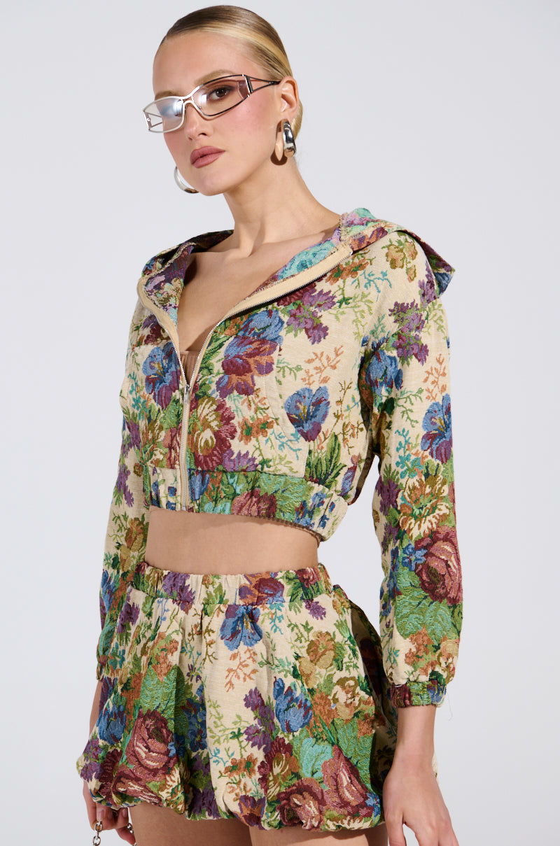 Donning the AKIRA Label ALL ABOUT ME FLORAL BOMBER JACKET and a matching skirt adorned with colorful flowers on a beige background exudes vintage charm. Paired with sunglasses and hoop earrings, their slicked-back hair enhances their confident pose against a plain backdrop.