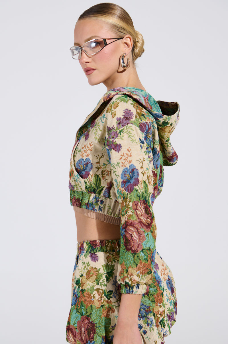 A blonde-haired individual with a bun wears clear glasses and an ALL ABOUT ME FLORAL BOMBER JACKET with matching shorts, showcasing vintage charm. The outfit's vibrant patterns stand out as they pose sideways against a plain background.