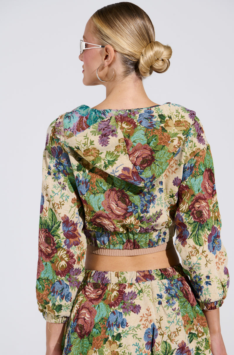 A person with blonde hair in a bun exudes vintage charm, wearing the colorful Floral Bomber Jacket from AKIRA Label's two-piece set. The top features a hood and cropped length, while they face away from the camera sporting large hoop earrings and sunglasses.