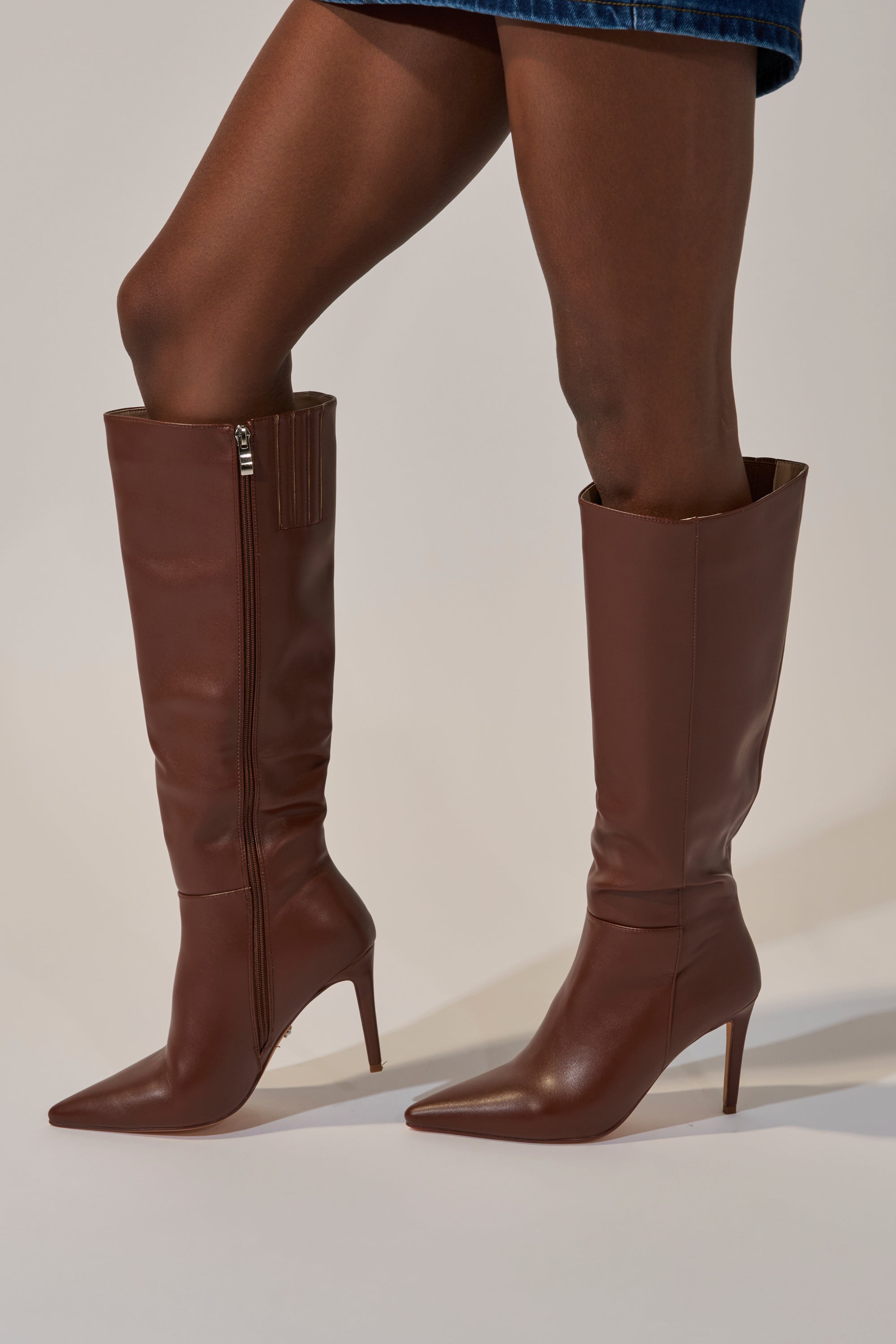 Someone wearing the AZALEA WANG HAILEYANN BROWN STILETO BOOT—knee-high, pointed toe, side zipper, brown faux leather with sleek fitted styling and elegant heels—paired with a blue skirt.