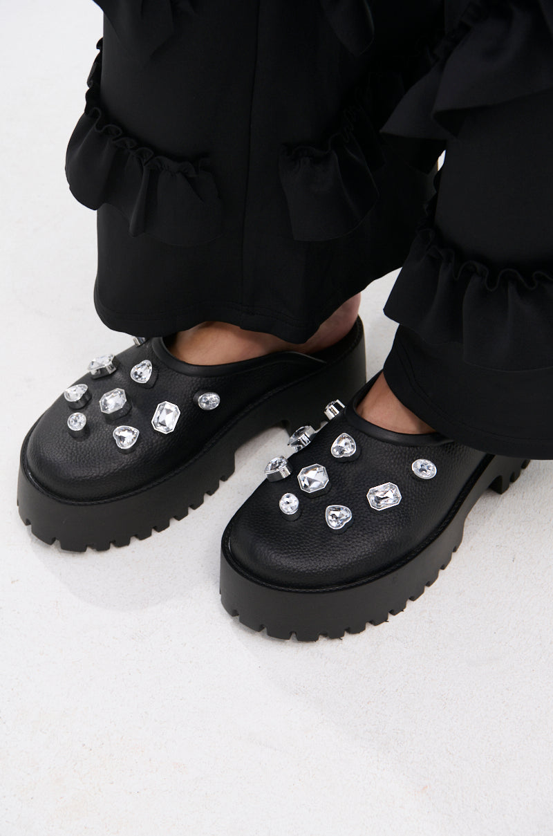 Close-up of AZALEA WANG HAIN BLACK EMBELLISHED FLAT shoes in faux leather with chunky heels and large clear gem accents, styled with black ruffled pants on a light floor.