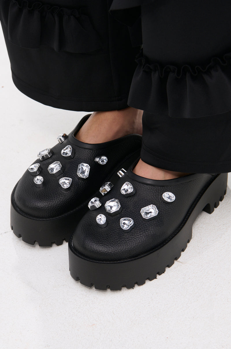 Close-up of the AZALEA WANG HAIN BLACK EMBELLISHED FLAT, a black faux leather platform shoe with chunky heels and large clear rhinestones. Worn with ruffled-hem black pants, shown on a light-colored floor.