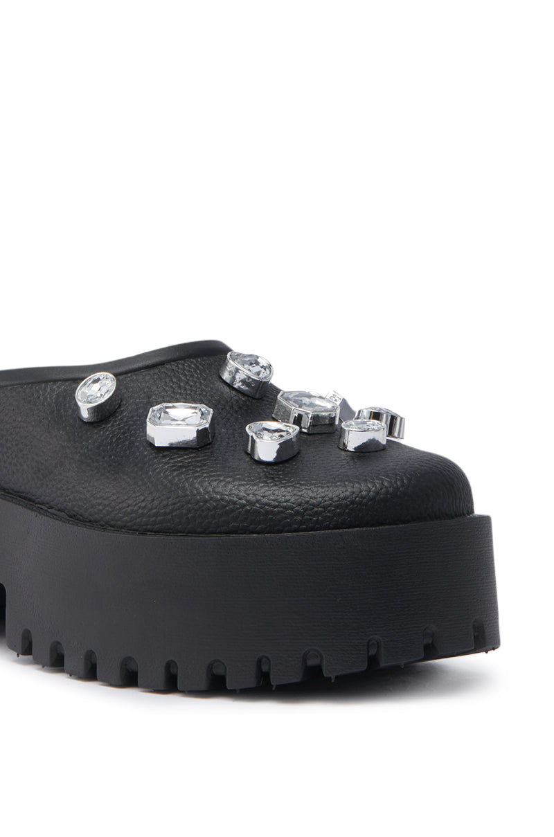 A close-up of the AZALEA WANG HAIN BLACK EMBELLISHED FLAT, a black faux leather platform clog with a rugged sole, chunky heel, and several large clear gemstones adorning the upper.