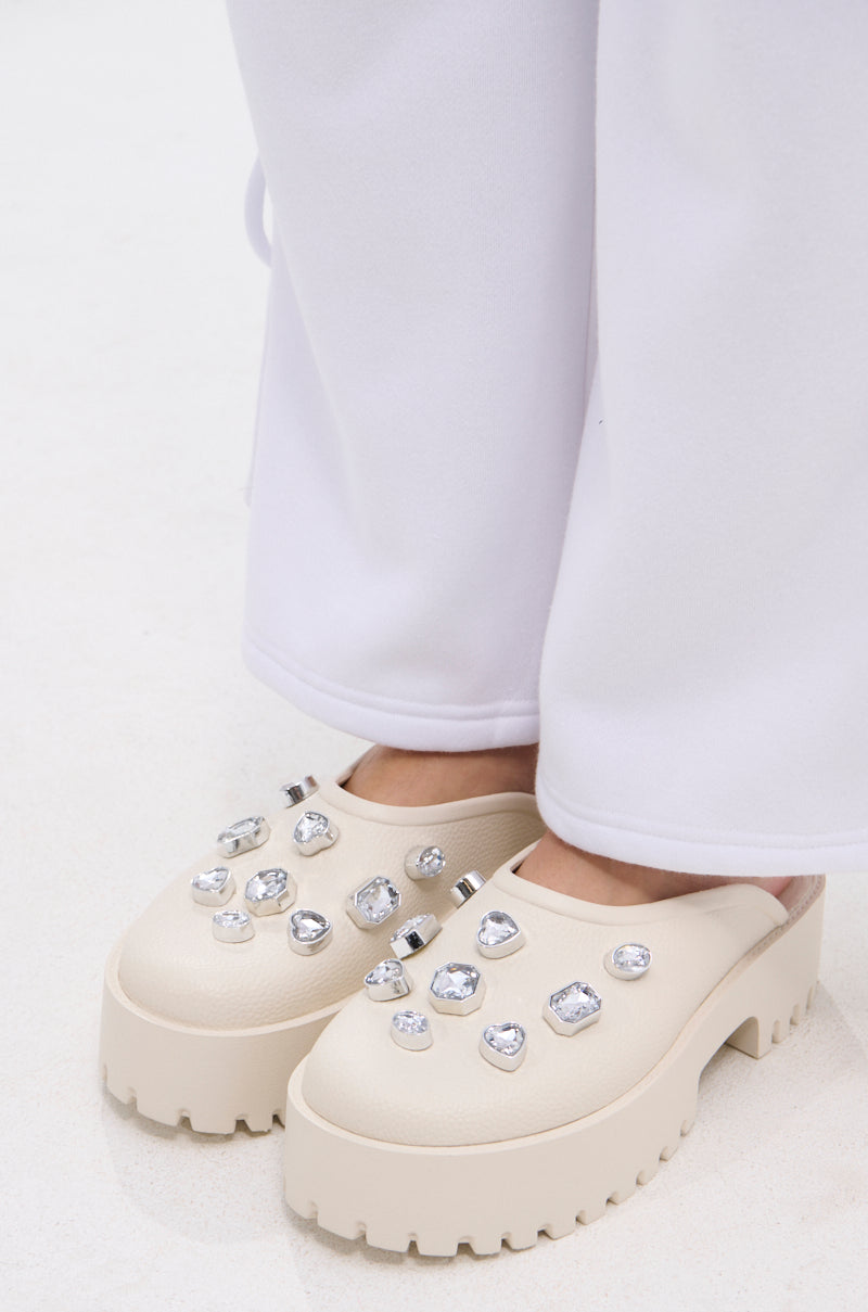 A person wears the AZALEA WANG HAIN IVORY RHINESTONE FLAT, chunky-heeled faux leather slip-ons with rhinestones. Loose white pants partially cover the dazzling flats in this close-up.