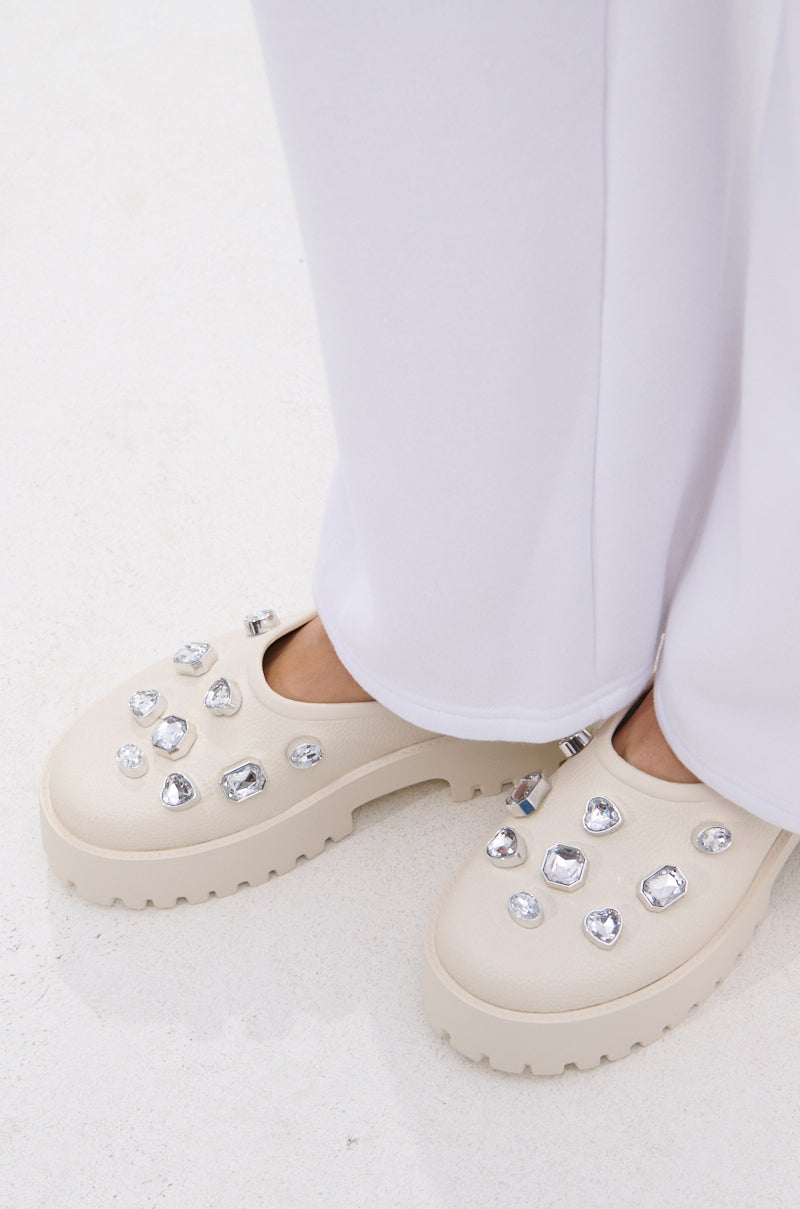 A person wears the AZALEA WANG HAIN IVORY RHINESTONE FLAT, featuring faux leather and sparkling rhinestones, standing on a white surface with long white pants nearly covering the chic, elegant shoes.