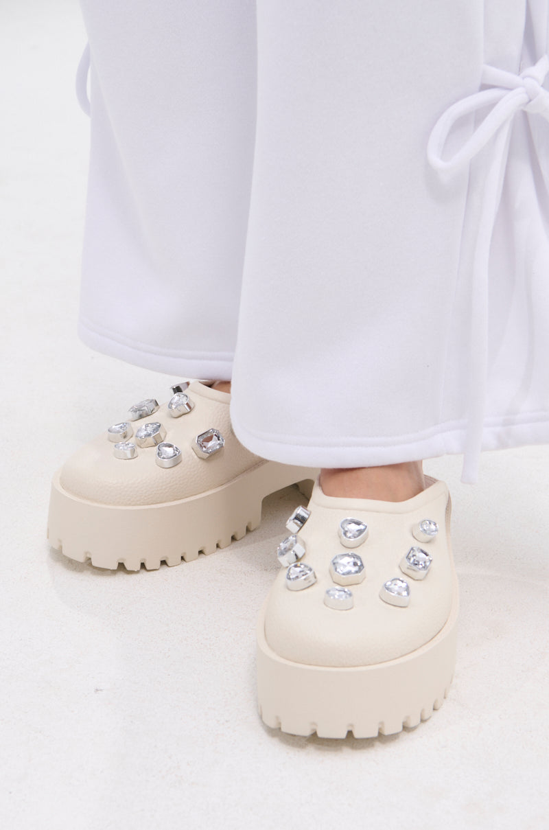 A person wearing wide-legged white pants with side ties and AZALEA WANG HAIN IVORY RHINESTONE FLAT shoes stands on a light-colored surface.