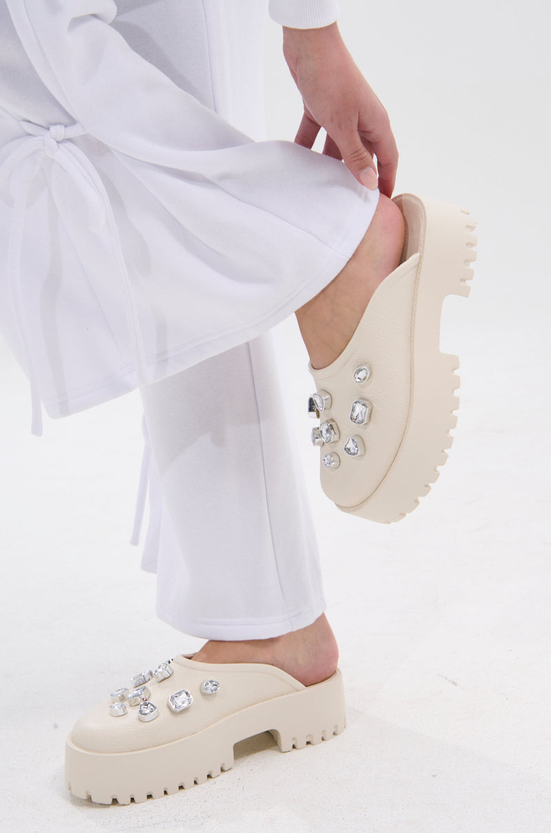 A person in white pants lifts one foot, showcasing the AZALEA WANG HAIN IVORY RHINESTONE FLAT—faux leather platform slip-ons adorned with large clear gemstones. The background is plain white.