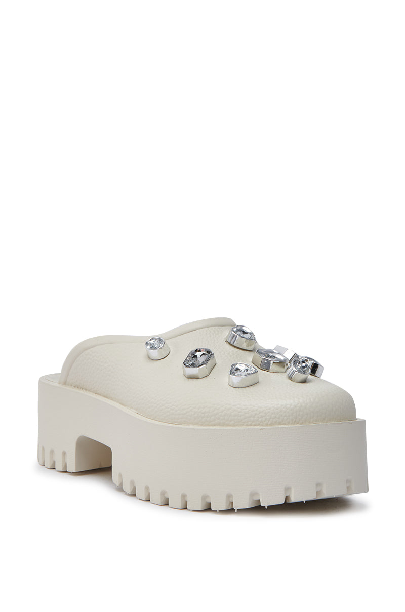 The AZALEA WANG HAIN IVORY RHINESTONE FLAT is a faux leather slip-on mule with a chunky textured sole and large clear rhinestones on the upper.
