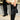 A person wearing the AZALEA WANG HALINA black croc stiletto boots, which feature a pointed toe silhouette, and holding a matching handbag stands on a street. The outfit includes a black and red top paired with beige shorts, set against the backdrop of a yellow barrier and pavement.