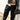 A person stands against a white wall, wearing a black leather jacket, high-waisted brown lace-up shorts, and AZALEA WANG HALINA BLACK SNAKE THIGH HIGH BOOTS, holding a small black handbag.