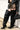 A person stands against a white wall, wearing a black leather jacket, high-waisted brown lace-up shorts, and AZALEA WANG HALINA BLACK SNAKE THIGH HIGH BOOTS, holding a small black handbag.