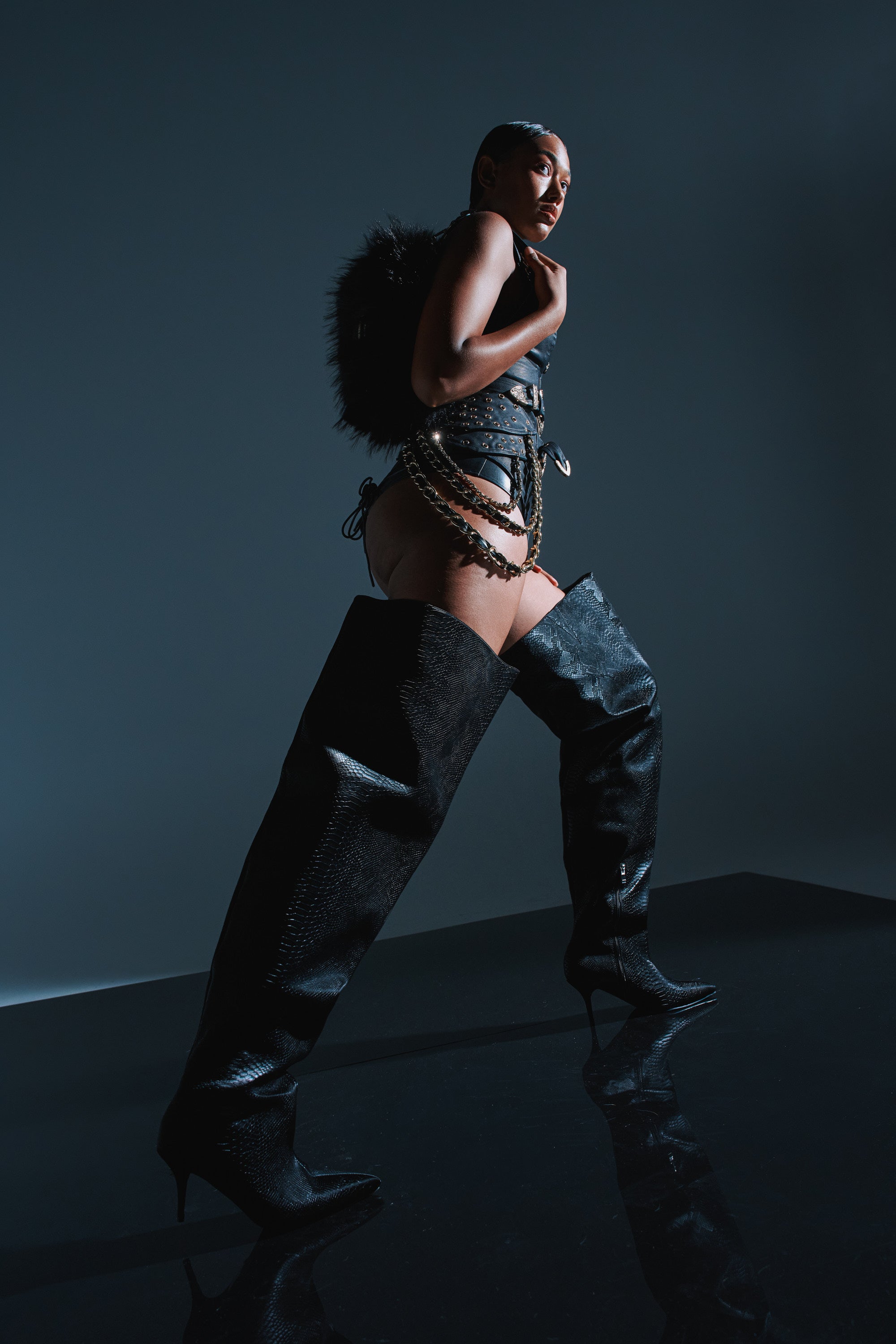 A person stands confidently on a glossy black surface, wearing a dramatic fashion outfit and the AZALEA WANG HALINA BLACK SNAKE THIGH HIGH BOOT, paired with strappy harnesses, chains, and a textured feathered accessory against a dark background.