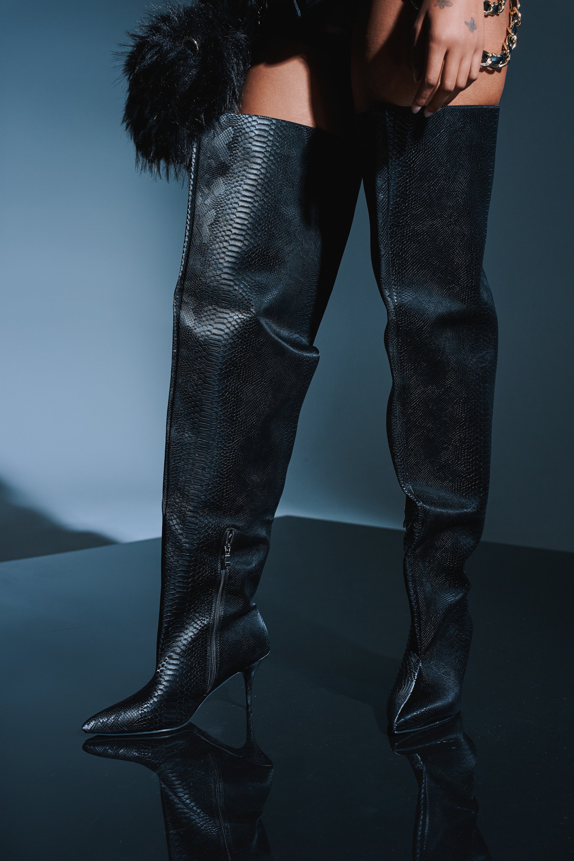 Only the lower body is visible as someone stands on a glossy black surface, wearing the AZALEA WANG HALINA BLACK SNAKE THIGH HIGH BOOT—a sleek, snake-print stiletto boot—against a dark background.