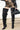 A person wearing AZALEA WANG HALINA BLACK SNAKE THIGH HIGH BOOTS, black and gold metallic shorts, a black leather jacket, and holding a small black handbag stands on the sidewalk.