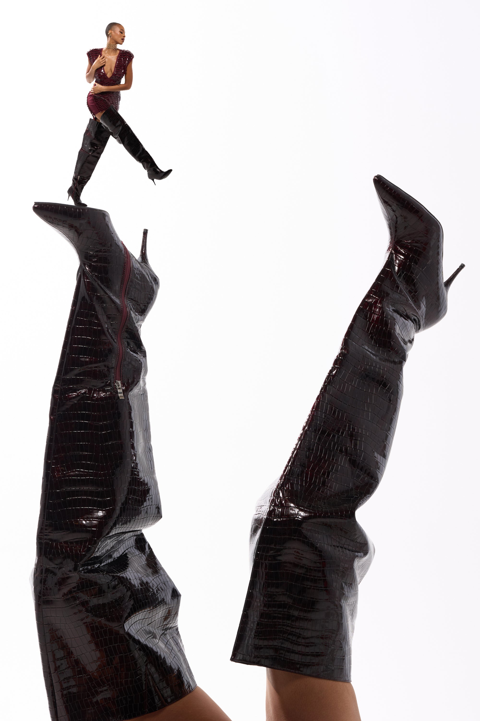 Wearing the AZALEA WANG HALINA BURGUNDY CROC STILETTO BOOT, a person lifts their legs as a tiny version of them balances on the tip, creating a surreal and playful scene against a white background. 0