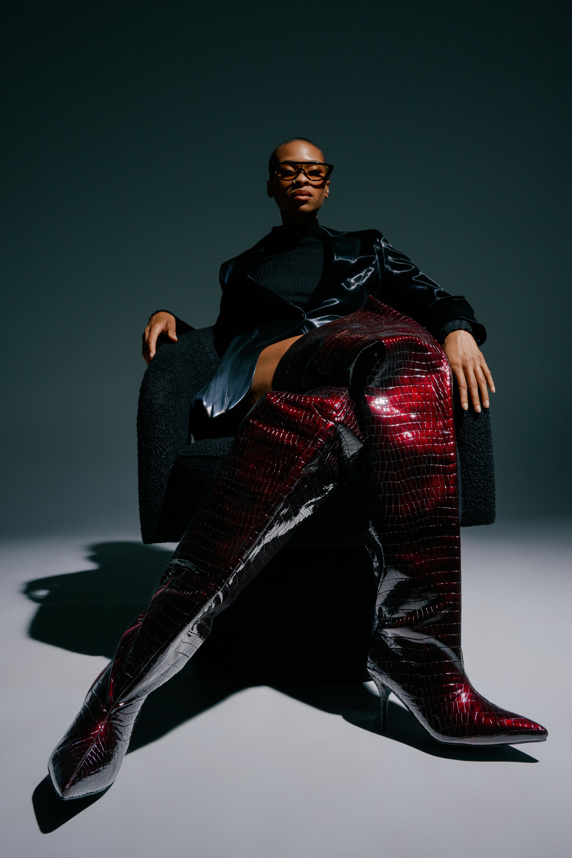 Outfitted in sleek black and confidently seated on a dark chair against a plain backdrop, the person showcases the AZALEA WANG HALINA BURGUNDY CROC STILETTO BOOT—striking burgundy thigh-highs that command attention.