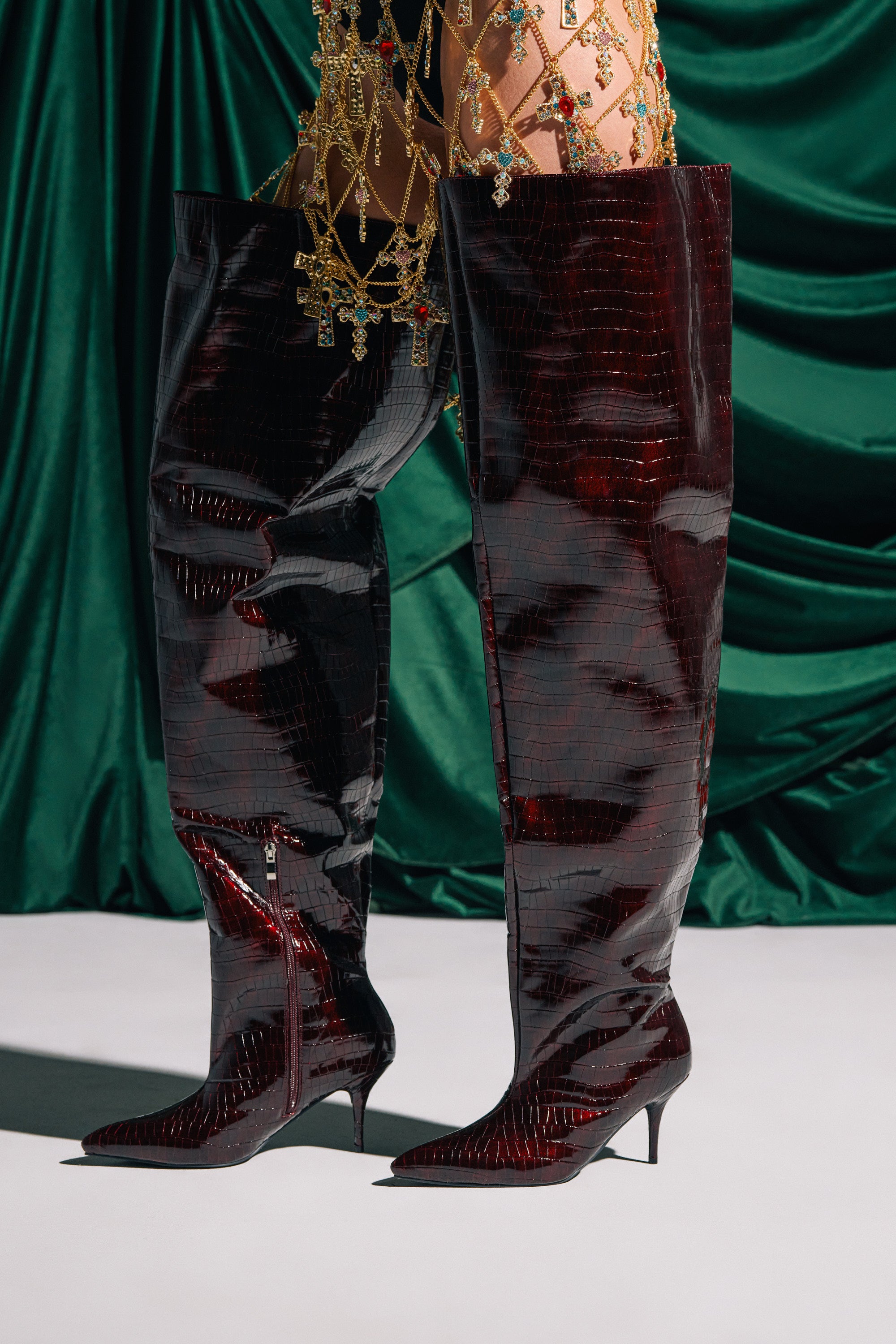 Wearing the AZALEA WANG HALINA BURGUNDY CROC STILETTO BOOT with a crocodile texture and gold chain accents, a person stands on a white floor before green draped fabric.