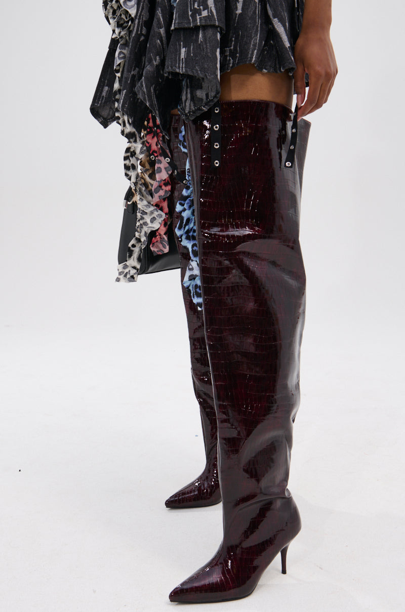 A person wears the AZALEA WANG HALINA BURGUNDY CROC STILETTO BOOT with pointed toes and kitten heels, styled with a ruffled monochrome skirt.
