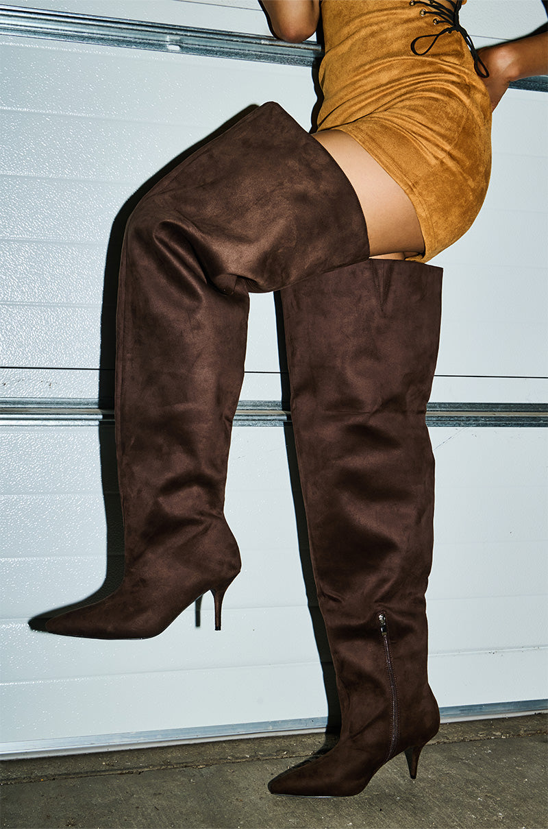 A person wearing a short tan dress and the AZALEA WANG HALINA CHOCOLATE THIGH HIGH BOOT with stiletto heels poses with one leg bent against a white garage door.