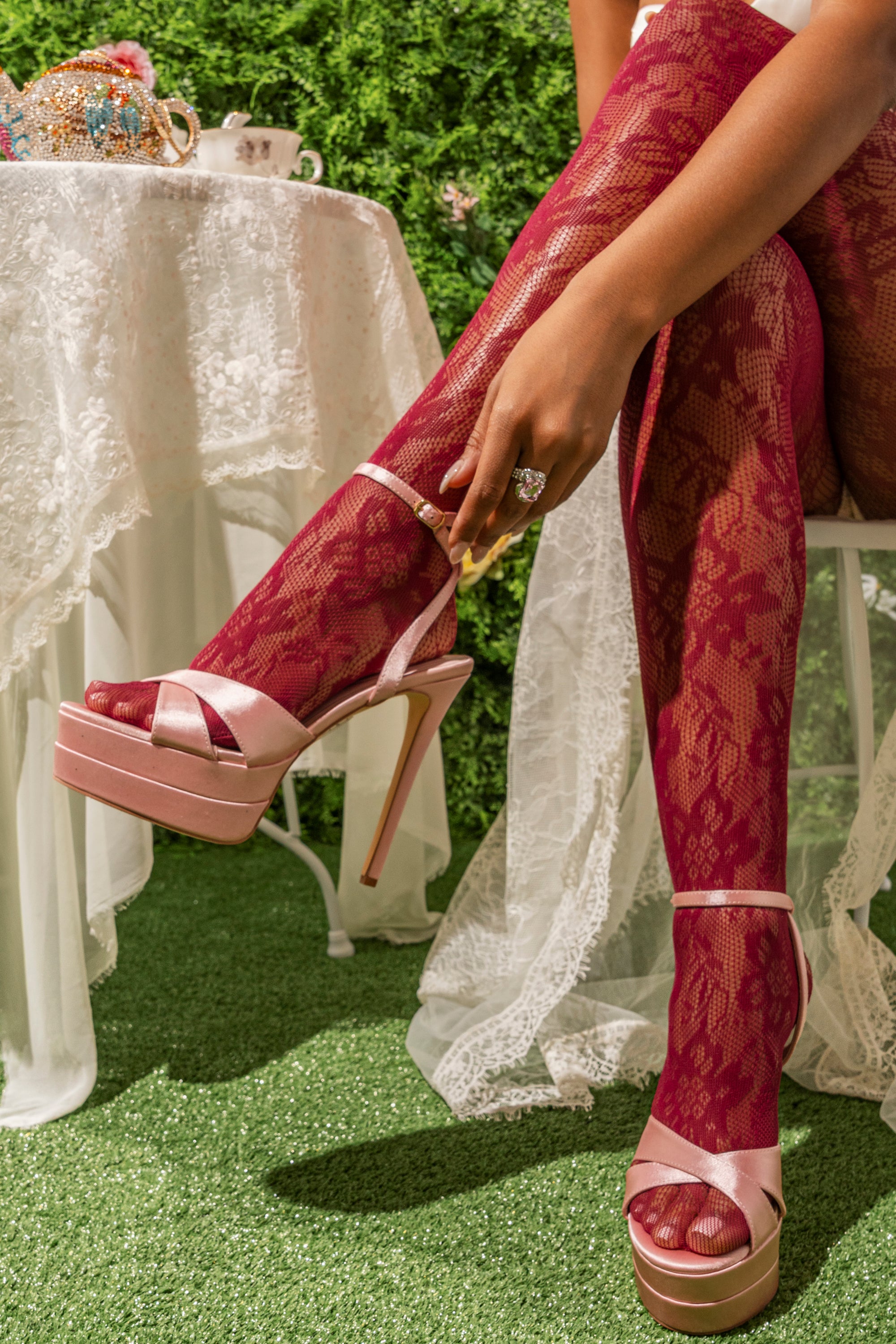 A person wearing pink lace tights and AZALEA WANG HALLADAY PINK SATIN PLATFORM STILETTO SANDALS sits at a lace-covered table outdoors on green grass, with a teapot and teacup on the table. 1