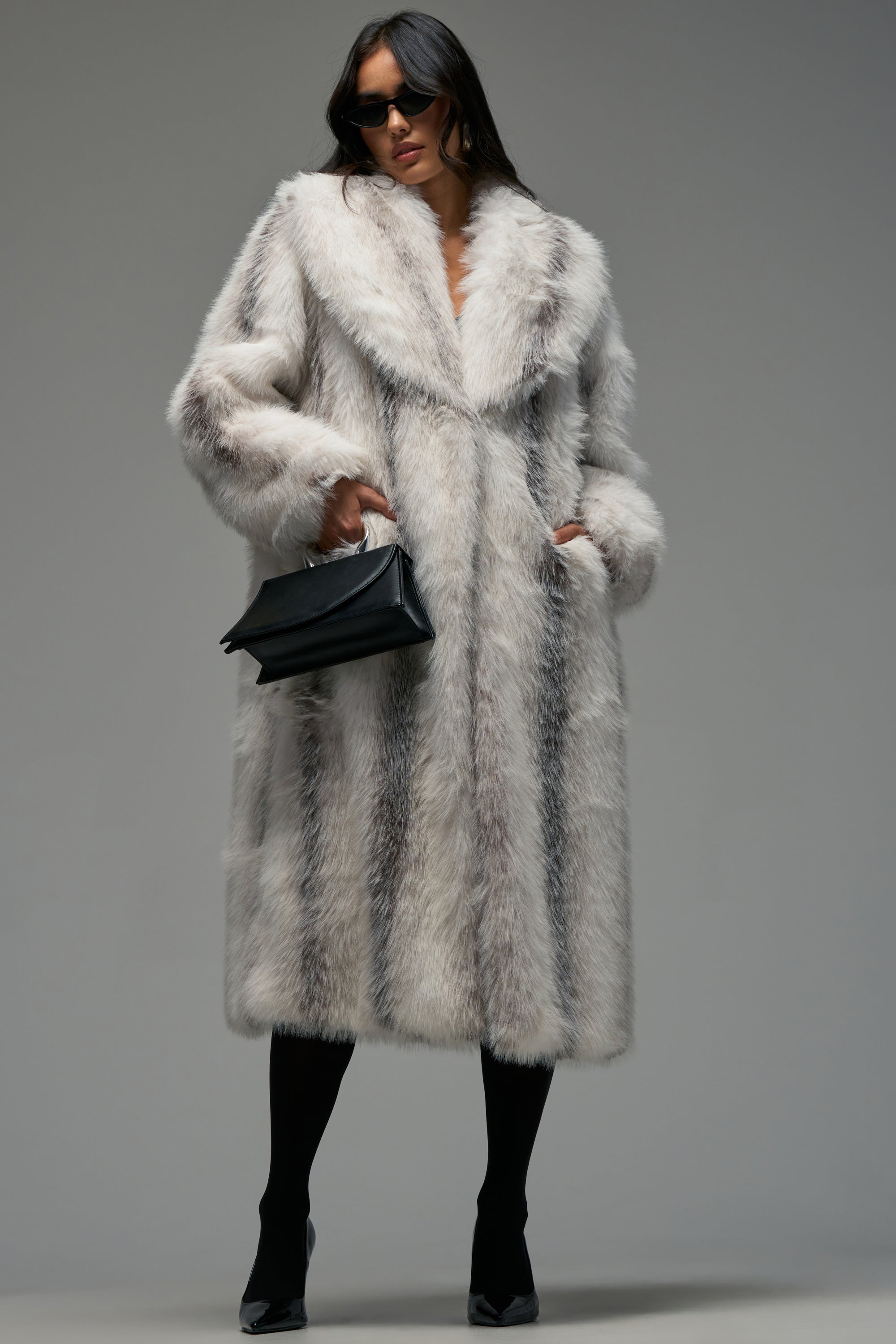 A person wearing the HALO MAXI FAUX FUR luxurious long winter coat with white and gray faux fur, paired with black tights, black shoes, sunglasses, and holding a black handbag stands against a plain gray background.
