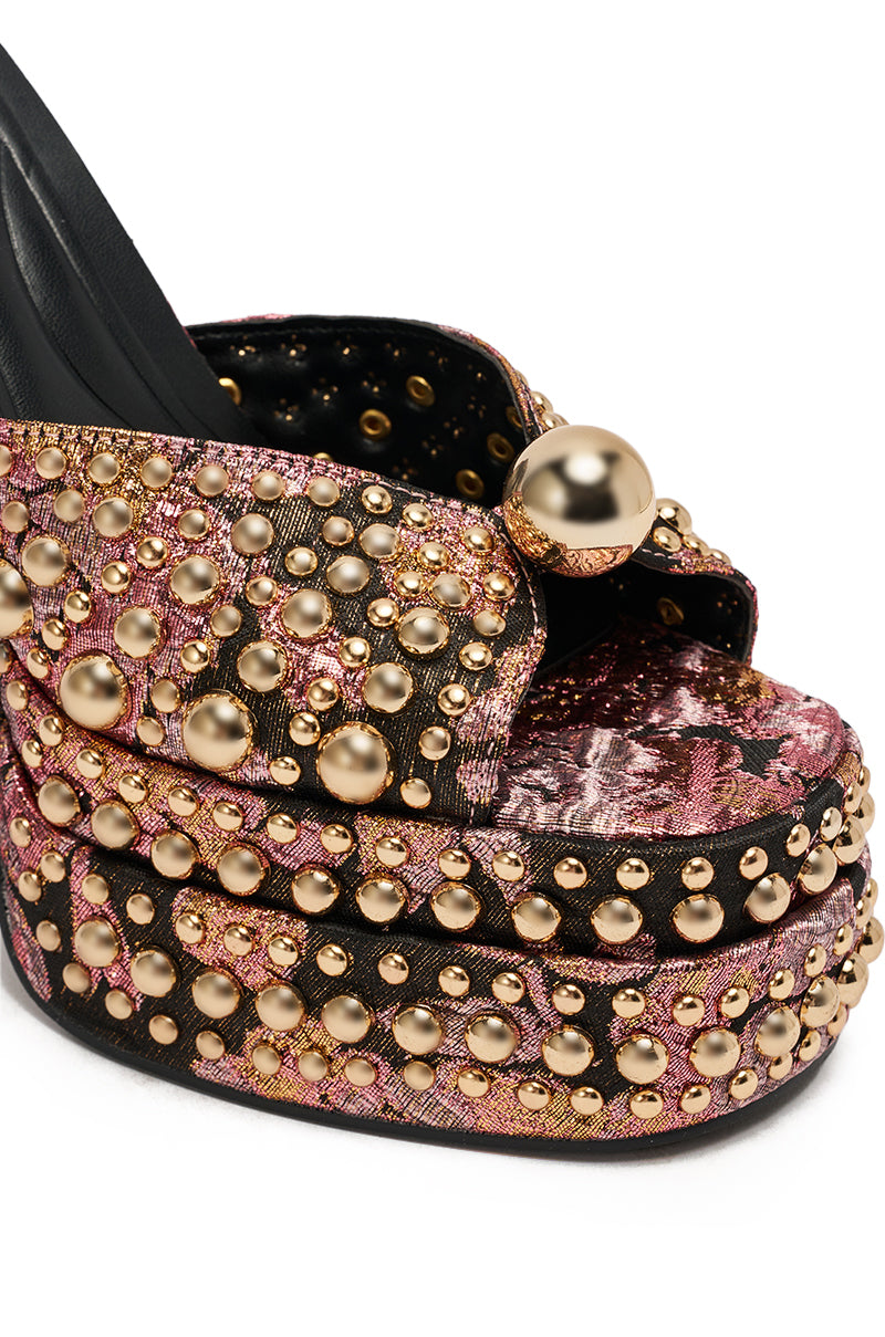 The AKIRA HANNESS BLACK MULTI BROCADE EMBELLISHED CHUNKY SANDAL boasts a high platform heel, black, pink, and gold brocade fabric, gold metallic studs, and a large gold sphere accent on crisscrossed straps.