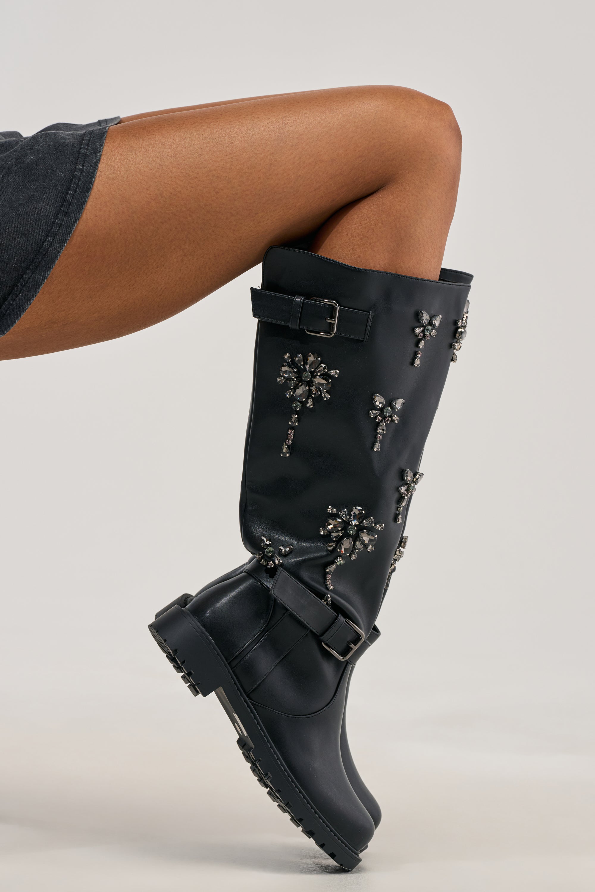 A close-up of a person’s leg wearing the AZALEA WANG HANOVER BLACK EMBELLISHED FLAT BOOT adorned with sparkling gemstone details, paired with a black skirt against a plain light background.