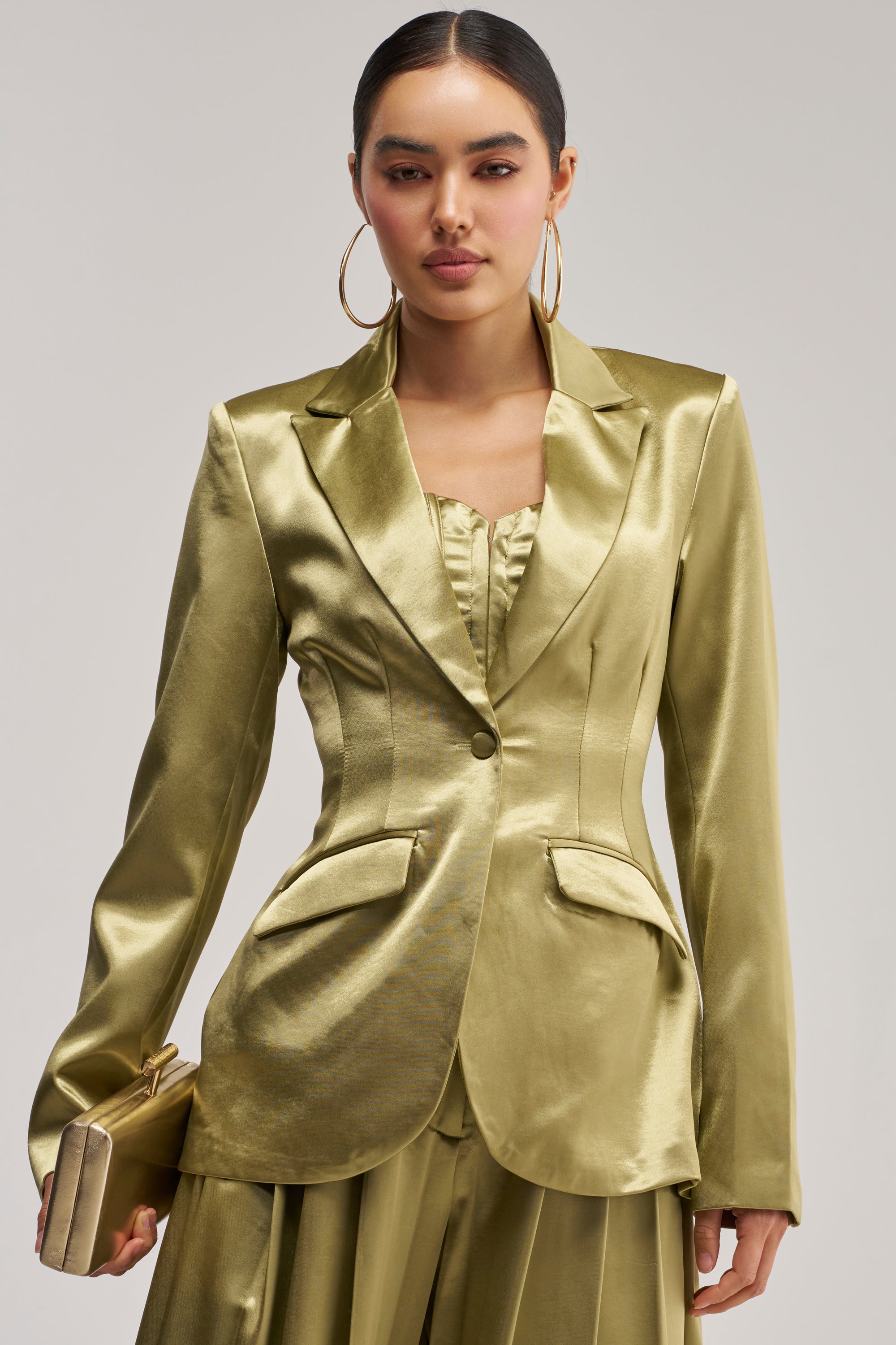 A woman wears the IRIS Satin Blazer in Olive, paired with a matching skirt and top, and holds a gold clutch. She accessorizes with hoop earrings, her sleek hair pulled back, and maintains a neutral expression against a plain light background.
