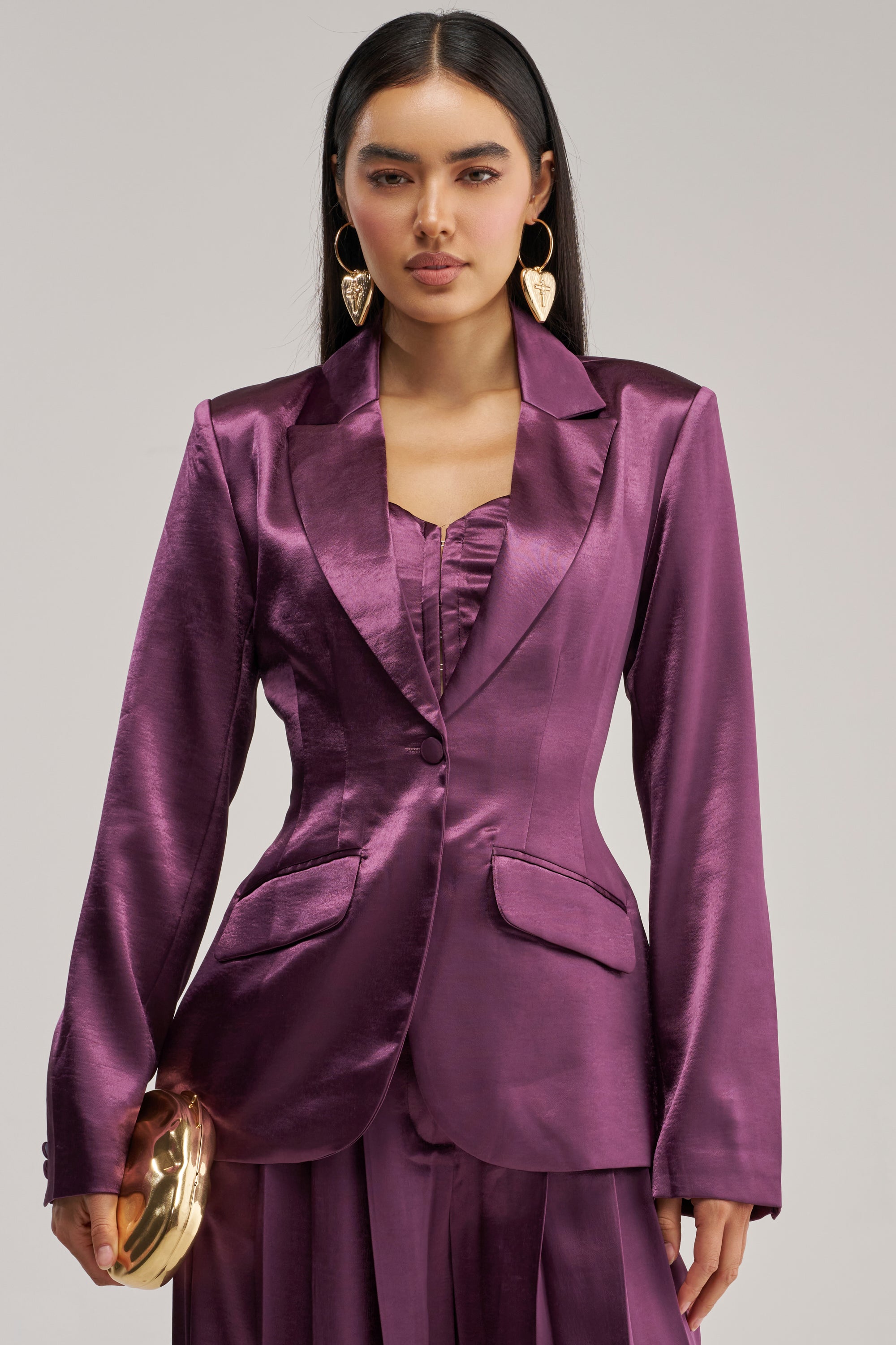 Wearing the IN TOO DEEP SATIN BLAZER in a lustrous plum satin, a woman with heart-shaped earrings holds a gold clutch while standing against a plain background.
