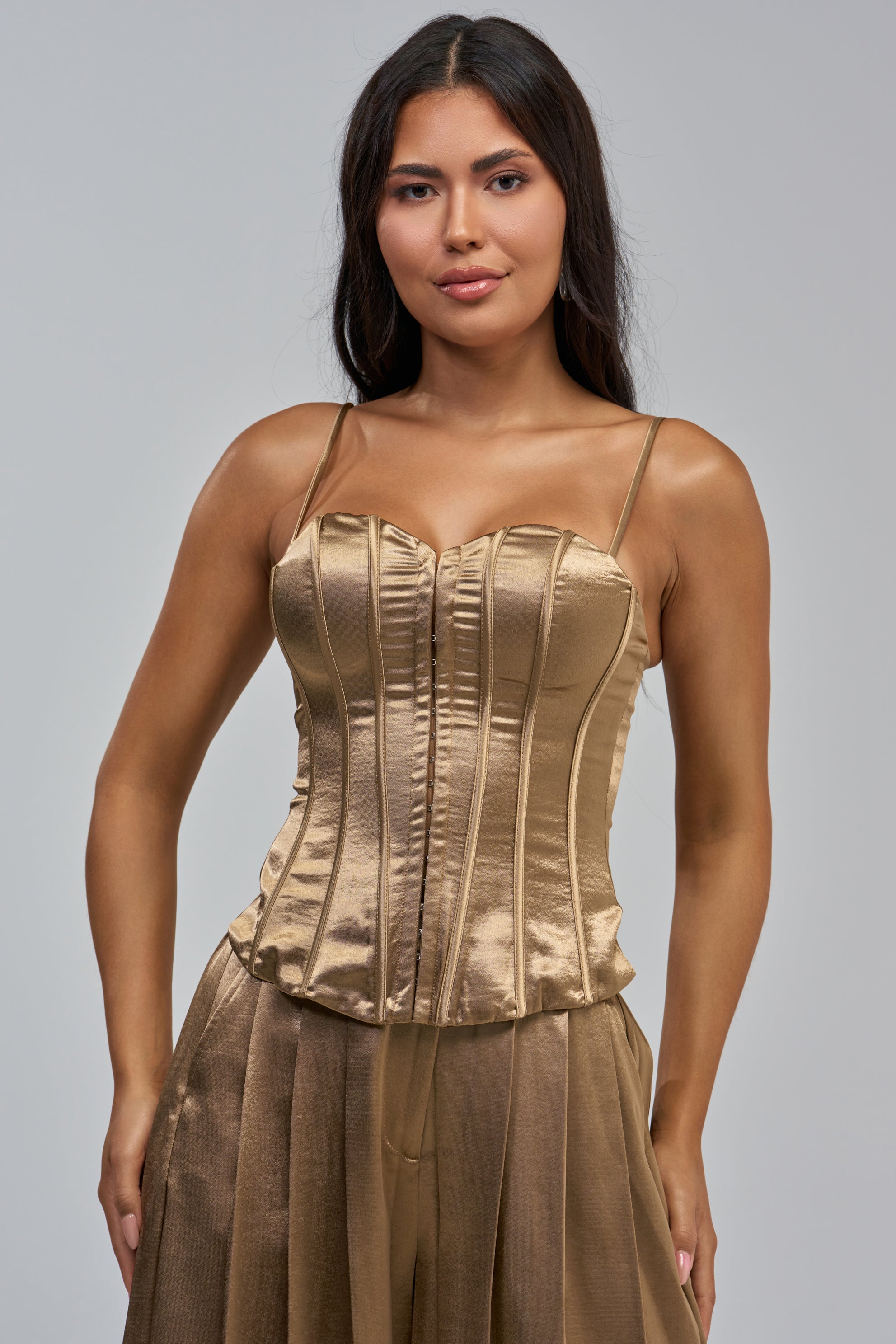 A woman with long, dark hair is wearing the IRIS SATIN CORSET IN LIGHT BROWN, which features structural boning and delicate spaghetti straps, paired with a matching pleated skirt—perfect for evening styling—as she poses against a plain grey background.