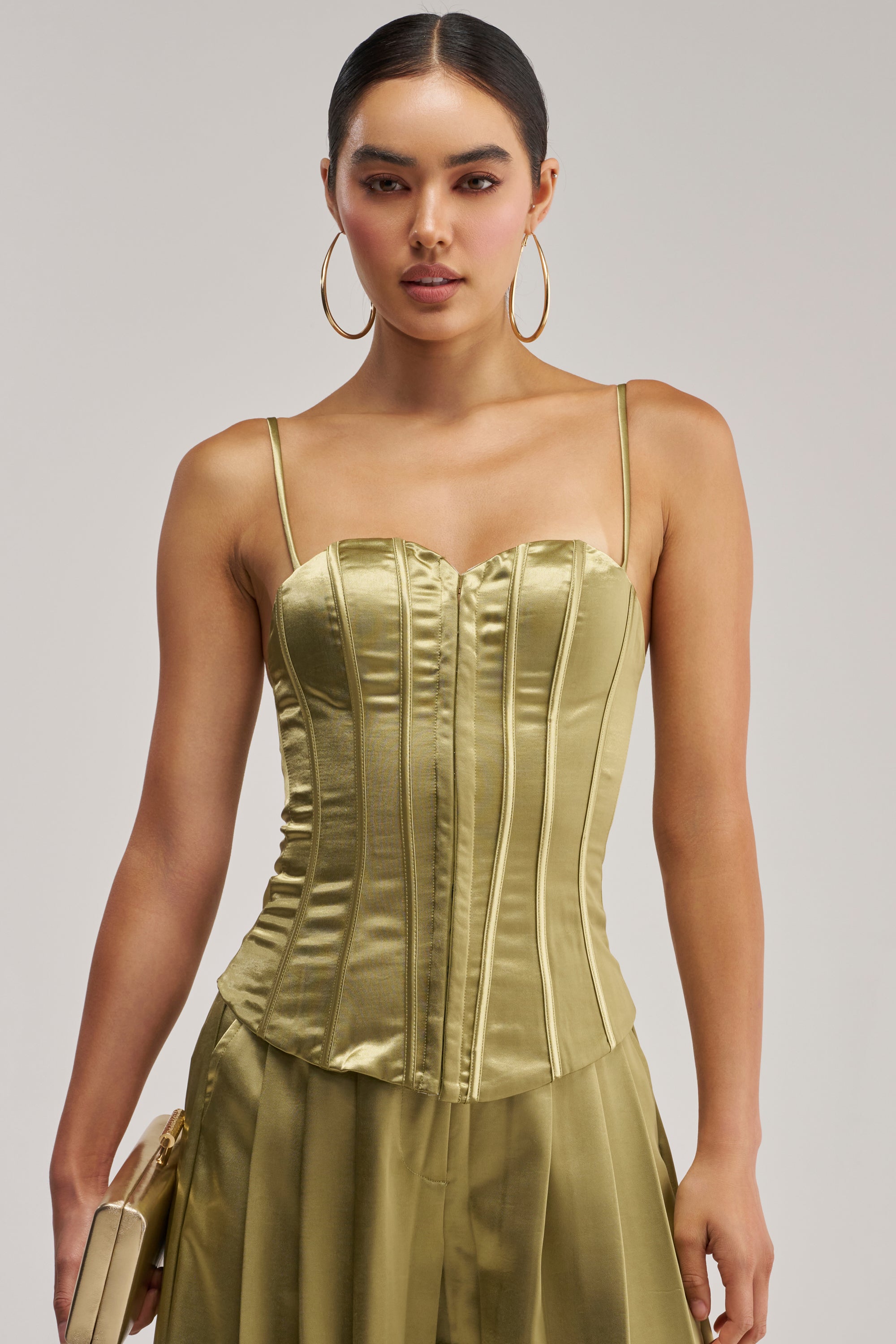 A woman wears the IRIS SATIN CORSET IN OLIVE paired with a coordinating skirt and gold clutch. With her hair slicked back, hoop earrings, and a neutral expression, she stands against a simple backdrop—an ideal choice for anyone seeking an elegant date night top.