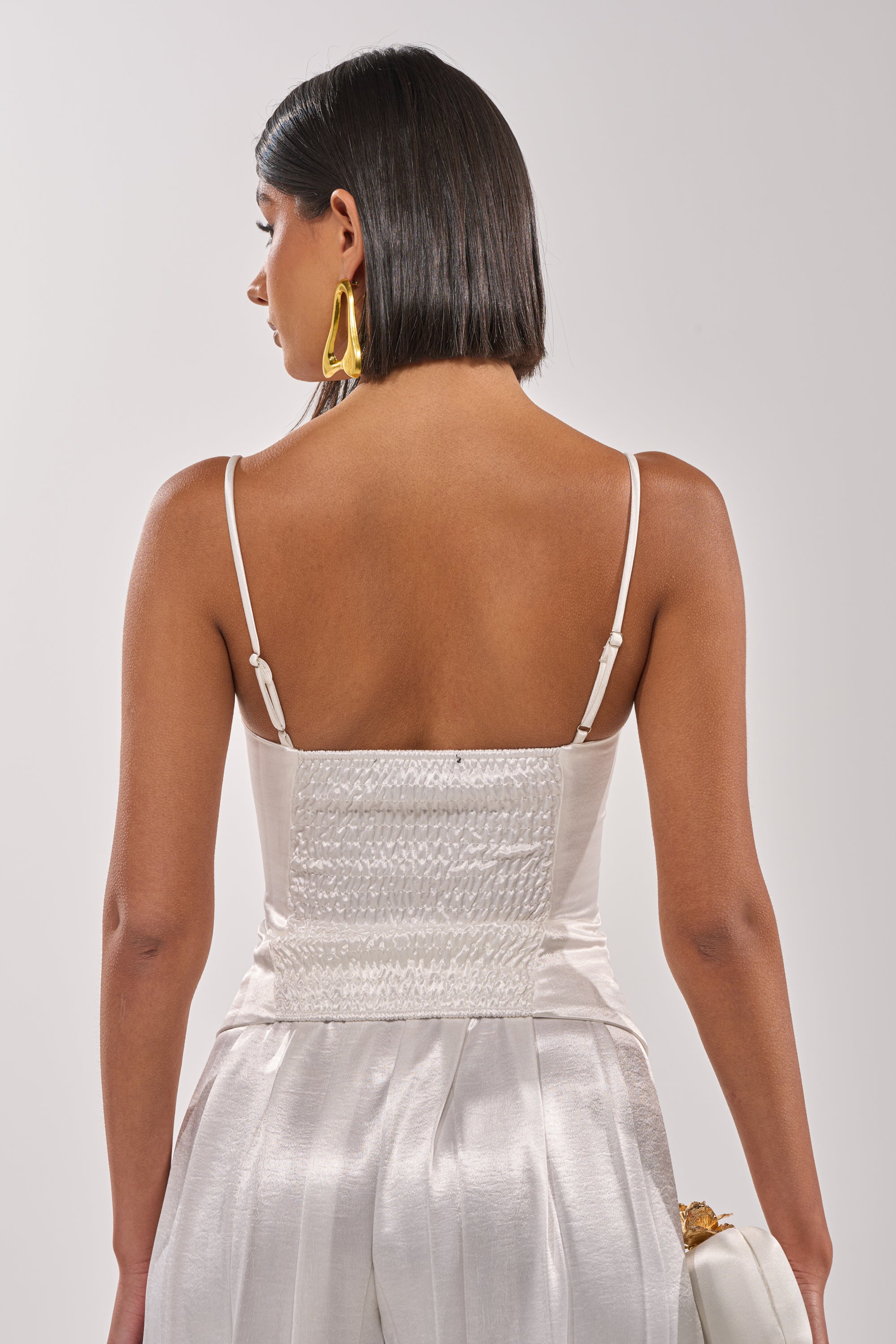 A woman with shoulder-length dark hair faces away, wearing the IRIS SATIN CORSET and pleated white pants. She accessorizes with a white purse and large gold earrings. 3